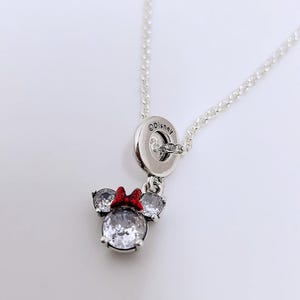 May include: Silver necklace with a Minnie Mouse pendant. The pendant has a large, clear crystal centre stone, two smaller side stones, and a red bow. The necklace has a fine chain and a Disney-branded clasp.