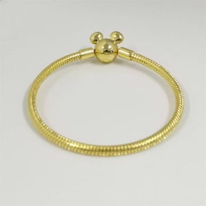 Pu&ograve; includere: Un braccialetto color oro con chiusura a forma di Mickey Mouse. Il braccialetto ha un design testurizzato a forma di serpente. La chiusura presenta un centro rotondo con due piccole orecchie rotonde sulla parte superiore. Su sfondo bianco.