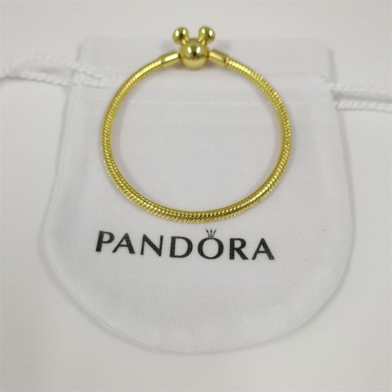 Pu&ograve; includere: Un braccialetto color oro con una chiusura a forma di Mickey Mouse &egrave; appoggiato su un sacchetto bianco. Il braccialetto ha un design strutturato a forma di serpente. Il sacchetto ha la scritta "PANDORA" stampata in nero.
