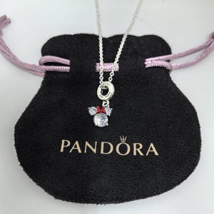 May include: A silver necklace with a Minnie Mouse pendant. The pendant has a large, clear crystal centre stone, two smaller side stones, and a red bow. The necklace is displayed on a black pouch with the "PANDORA" logo.