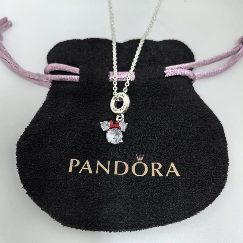May include: A silver necklace with a Minnie Mouse pendant. The pendant has a large, clear crystal centre stone, two smaller side stones, and a red bow. The necklace is displayed on a black pouch with the "PANDORA" logo.