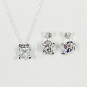 May include: A silver necklace and matching earrings set. The pendant features a square, clear gemstone set within a Stitch-shaped frame with red, blue, and pink accents. The earrings mirror the design, showcasing the same character in a smaller size.