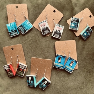 May include: A collection of book-shaped earrings. The earrings display titles such as "Ugly Love," "Never Lie," and "Housemaid." Each miniature book has a detailed cover design and is suspended from a silver earring hook.