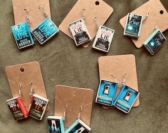 May include: Earrings featuring miniature book designs. The earrings showcase various book titles, including "Ugly Love," "Never Lie," and "Housemaid." Each book has a detailed cover design and is attached to a silver earring hook.