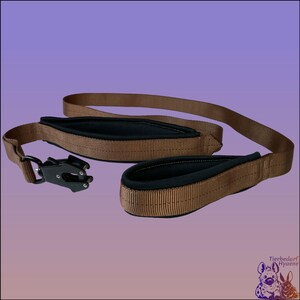 Pode incluir: Uma trela castanha para cães com pegas almofadadas pretas e um fecho de metal preto. A trela é feita de material durável e foi concebida para conforto e controlo durante as caminhadas. A trela é mostrada sobre um fundo roxo.