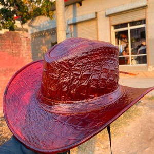 Peut inclure: Un chapeau de cowboy rouge fonc&eacute; pr&eacute;sentant une texture en relief imitant la peau de crocodile sur toute sa surface. Le chapeau poss&egrave;de une calotte structur&eacute;e, un large bord incurv&eacute; et une fine mentonni&egrave;re noire fix&eacute;e &agrave; la base.