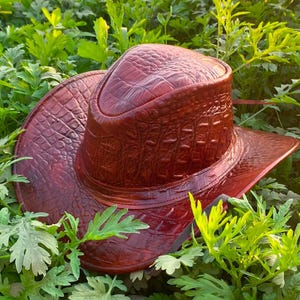 Peut inclure: Un chapeau de cowboy brun rouge&acirc;tre profond pr&eacute;sentant une texture en relief fa&ccedil;on peau de crocodile sur toute la surface, y compris la calotte et le bord large et incurv&eacute;, avec une finition brillante et des motifs d'&eacute;cailles d&eacute;taill&eacute;s.