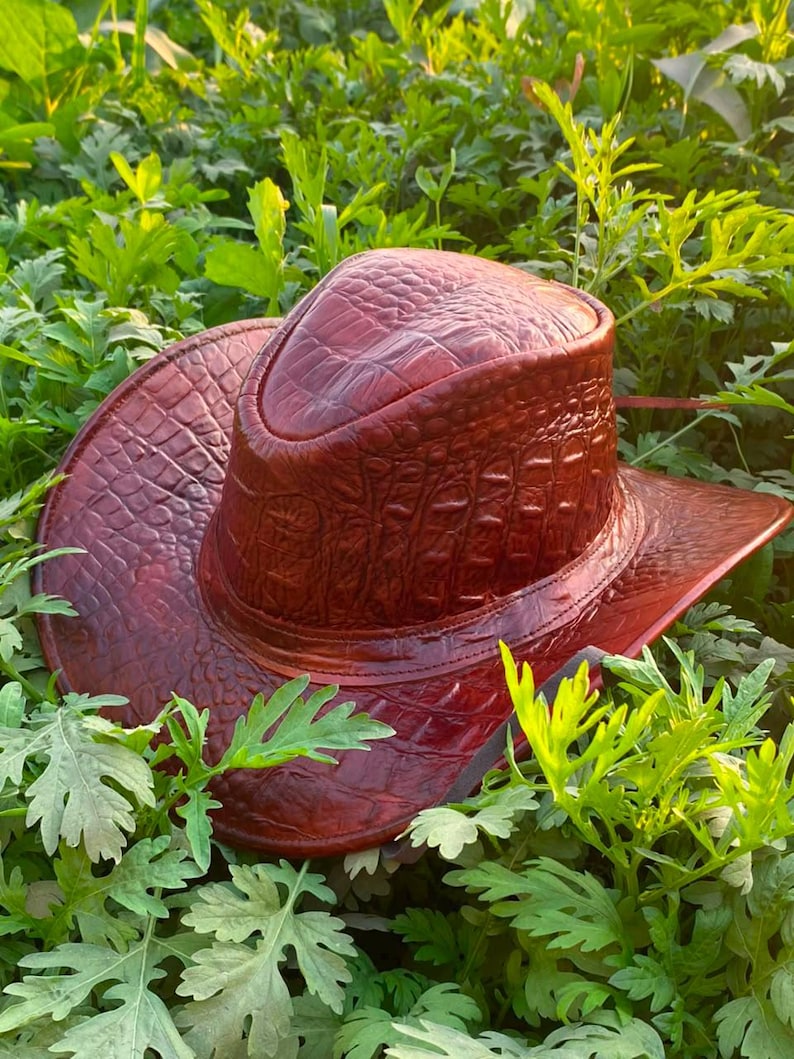 Peut inclure: Un chapeau de cowboy brun rouge&acirc;tre profond pr&eacute;sentant une texture en relief fa&ccedil;on peau de crocodile sur toute la surface, y compris la calotte et le bord large et incurv&eacute;, avec une finition brillante et des motifs d'&eacute;cailles d&eacute;taill&eacute;s.