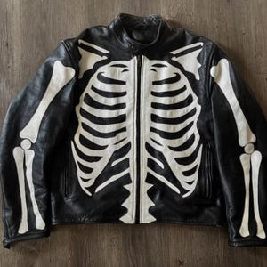May include: A black leather jacket featuring a white skeleton bone graphic across the front and sleeves. The design includes a rib cage pattern on the torso, shoulder blade details, and arm bones extending down the sleeves with a central zip.