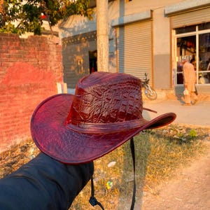 Peut inclure: Un chapeau de cowboy rouge fonc&eacute; pr&eacute;sentant un motif textur&eacute; en relief fa&ccedil;on peau de crocodile sur la calotte et les larges bords. Le chapeau comprend une fine jugulaire noire r&eacute;glable fix&eacute;e sur les c&ocirc;t&eacute;s pour maintenir le couvre-chef.