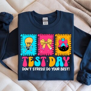 Pu&ograve; includere: Felpa blu navy con una grafica colorata "TEST DAY" in un font retr&ograve;. Il design include tre immagini incorniciate: una lampadina, un fiocco e un flacone di profumo. &Egrave; incluso anche il testo "DON'T STRESS! DO YOUR BEST!"