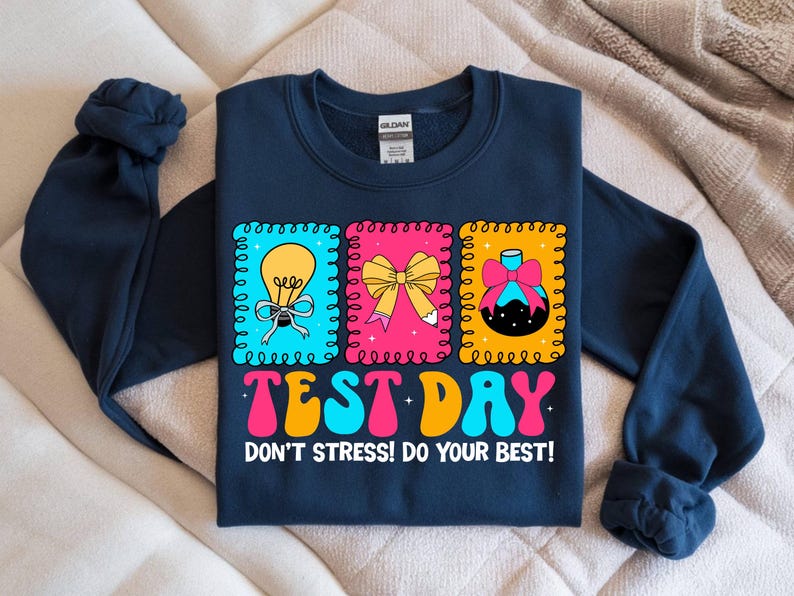 Pu&ograve; includere: Felpa blu navy con una grafica colorata "TEST DAY" in un font retr&ograve;. Il design include tre immagini incorniciate: una lampadina, un fiocco e un flacone di profumo. &Egrave; incluso anche il testo "DON'T STRESS! DO YOUR BEST!"