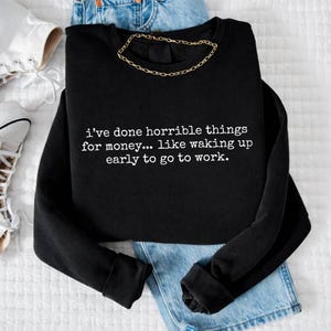 Puede incluir: Sudadera negra de manga larga con el texto "i've done horrible things for money... like waking up early to go to work." Una cadena de oro está sobre la sudadera. La sudadera está sobre unos vaqueros azules.