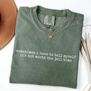May include: An olive green t-shirt with the text "sometimes i have to tell myself it's not worth the jail time" in white. A gold necklace with a pendant rests on the shirt. A brown hat and dried plant are in the background.