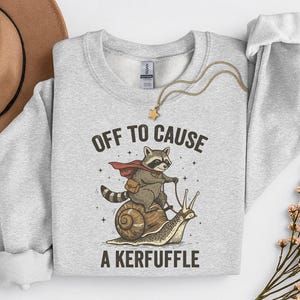 May include: Light grey sweatshirt with a graphic of a raccoon riding a snail, wearing a cape and satchel. Text reads "OFF TO CAUSE A KERFUFFLE". A gold necklace with a star pendant and a brown hat are also visible.