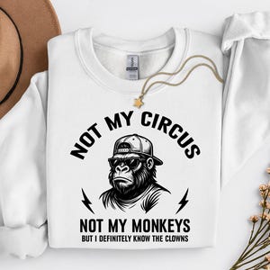 May include: White sweatshirt with a graphic of a gorilla wearing sunglasses and a baseball cap. The text reads "NOT MY CIRCUS, NOT MY MONKEYS, BUT I DEFINITELY KNOW THE CLOWNS". A gold necklace with a star pendant is draped on the sweatshirt.
