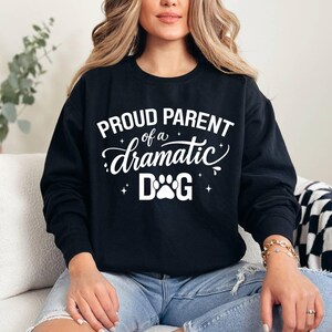 May include: Black sweatshirt with the text "PROUD PARENT of a dramatic DOG" in white script and a paw print graphic. The sweatshirt is a crew neck style and is made of a soft material. The design is centered on the front of the shirt.