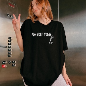 May include: Black t-shirt with the text "NO GOLF TODAY." in white, along with a stick figure. The shirt is worn by a person giving a peace sign. The person is standing in front of an elevator.