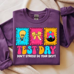 Pu&ograve; includere: Una felpa viola con un design grafico colorato. Il design presenta tre cornici rettangolari con una lampadina, un fiocco e un oggetto decorativo. Sono visualizzate anche le parole "TEST DAY" e "DON'T STRESS! DO YOUR BEST!"