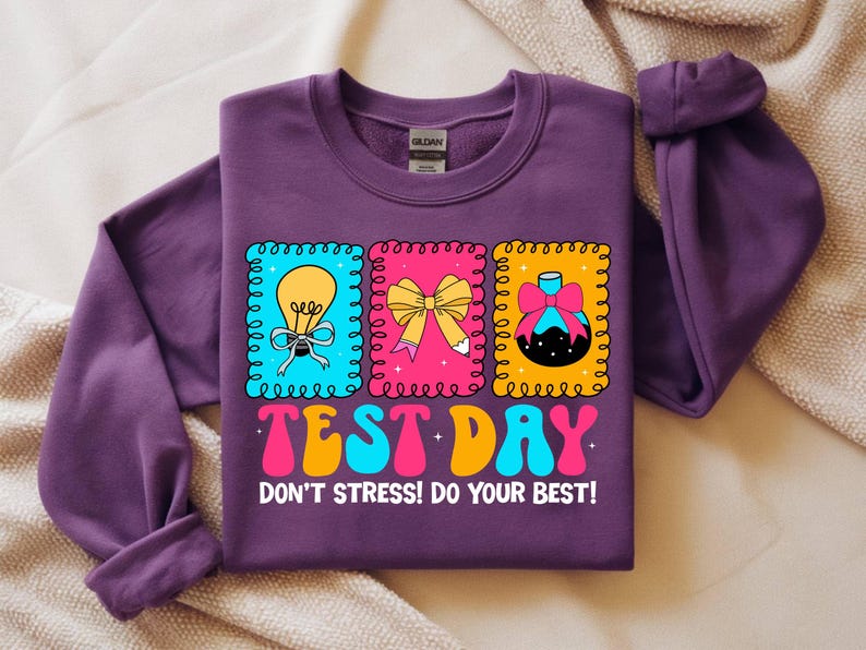 Pu&ograve; includere: Una felpa viola con un design grafico colorato. Il design presenta tre cornici rettangolari con una lampadina, un fiocco e un oggetto decorativo. Sono visualizzate anche le parole "TEST DAY" e "DON'T STRESS! DO YOUR BEST!"