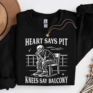 Puede incluir: Sudadera negra con un gráfico blanco de un esqueleto sentado en una silla, con el texto "HEART SAYS PIT KNEES SAY BALCONY". Un collar dorado con un colgante de estrella está sobre la sudadera.