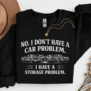 May include: Black sweatshirt featuring the text "NO, I DON'T HAVE A CAR PROBLEM. I HAVE A STORAGE PROBLEM." The design includes illustrations of four classic cars. A gold necklace with a star pendant is also visible.