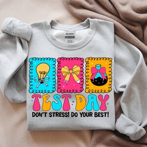 Pu&ograve; includere: Una felpa grigio chiaro con un design grafico colorato. Il design include tre immagini incorniciate di una lampadina, un fiocco e un ornamento decorativo. Il testo "TEST DAY" e "DON'T STRESS! DO YOUR BEST!" sono stampati.