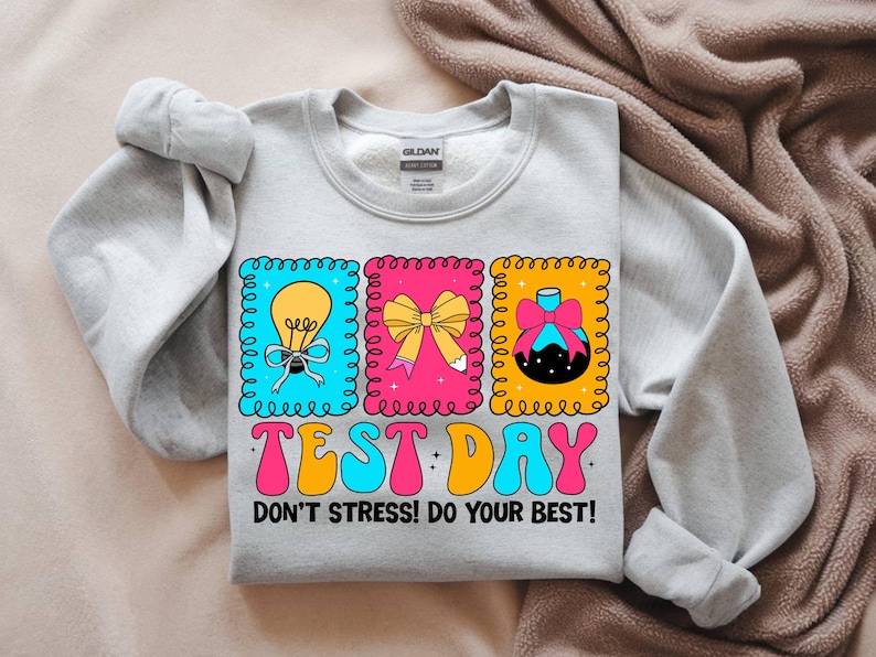 Pu&ograve; includere: Una felpa grigio chiaro con un design grafico colorato. Il design include tre immagini incorniciate di una lampadina, un fiocco e un ornamento decorativo. Il testo "TEST DAY" e "DON'T STRESS! DO YOUR BEST!" sono stampati.