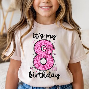 Op de afbeelding: Een lichtroze t-shirt met de tekst "it's my 8th birthday". Het cijfer 8 is in roze luipaardprint met een roze strik. Kleine roze hartjes en sterren staan ook op het shirt.
