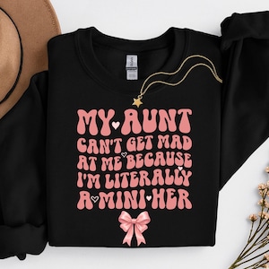 May include: Black sweatshirt with the text "MY AUNT CAN'T GET MAD AT ME BECAUSE I'M LITERALLY A MINI HER" in pink. A gold necklace with a star pendant and a pink bow are also visible.