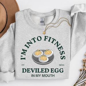 Op de afbeelding: Een lichtgrijs sweatshirt met de tekst "I'M INTO FITNESS DEVILED EGG IN MY MOUTH" in het groen. De afbeelding toont drie gevulde eieren op een bord. Een gouden ketting met een sterhanger en een bruine hoed staan ook op de foto.