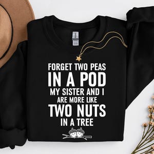 May include: Black crewneck sweatshirt with the text "FORGET TWO PEAS IN A POD MY SISTER AND I ARE MORE LIKE TWO NUTS IN A TREE" in white. A gold necklace with a star pendant is draped on the sweatshirt. A brown hat and floral arrangement are in the background.