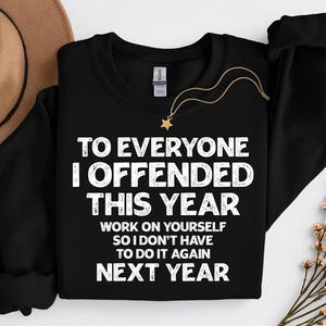 May include: Black crewneck sweatshirt with white text: "TO EVERYONE I OFFENDED THIS YEAR WORK ON YOURSELF SO I DON'T HAVE TO DO IT AGAIN NEXT YEAR." A gold necklace with a star pendant is draped on the sweatshirt. A brown hat and flowers are in the background.
