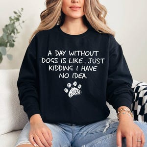 Puede incluir: Sudadera negra con texto blanco: "Un día sin perros es como... es broma, no tengo ni idea". Una huella de pata blanca está debajo del texto. La sudadera la lleva una persona.