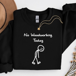 Op de afbeelding: Zwart sweatshirt met ronde hals met de tekst "No Woodworking Today" en een trieste stokfiguur. Een gouden ketting met een sterhanger ligt op de trui. Een bruine hoed en bloemen zijn op de achtergrond.