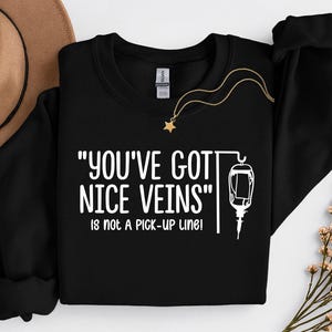 Puede incluir: Sudadera negra con texto blanco que dice "YOU'VE GOT NICE VEINS" y "IS NOT A PICK-UP LINE!". Una ilustración blanca de una bolsa intravenosa está debajo del texto. Un collar dorado con un colgante de estrella está sobre la sudadera.