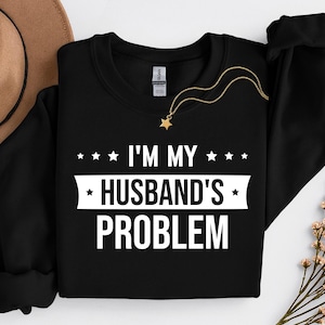 May include: Black sweatshirt with the text "I'M MY HUSBAND'S PROBLEM" in white. The sweatshirt has a crew neck and long sleeves. A gold necklace with a star pendant is draped on the sweatshirt. A brown hat and flowers are in the background.