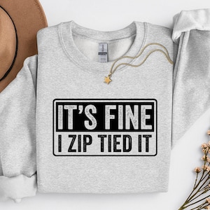 Puede incluir: Una sudadera gris claro con el texto "IT'S FINE I ZIP TIED IT" en un rectángulo negro. También se ve un collar dorado con un colgante de estrella y un sombrero marrón. La sudadera tiene cuello redondo y mangas largas.