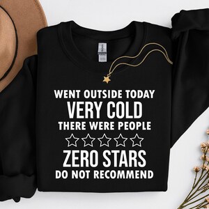 以下が含まれることがあります： 白い文字で「WENT OUTSIDE TODAY VERY COLD THERE WERE PEOPLE ZERO STARS DO NOT RECOMMEND」と書かれた黒いクルーネックのスウェットシャツ。星のペンダントが付いた金のネックレスがスウェットシャツの上にあります。茶色の帽子と花が背景にあります。