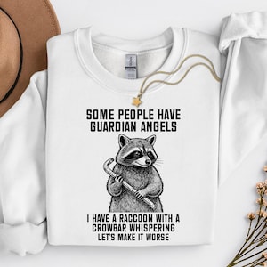 Puede incluir: Sudadera blanca con el texto "SOME PEOPLE HAVE GUARDIAN ANGELS. I HAVE A RACCOON WITH A CROWBAR WHISPERING LET'S MAKE IT WORSE." Un gráfico de mapache sosteniendo una palanca está impreso. Se ve un collar dorado.