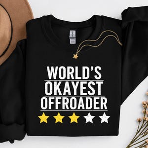 Könnte beinhalten: Schwarzes Sweatshirt mit dem weißen Text "WORLD'S OKAYEST OFFROADER". Das Design umfasst vier gelbe Sterne und einen weißen Stern. Eine goldene Halskette mit Sternanhänger liegt auf dem Sweatshirt.