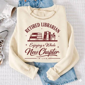 May include: Cream-colored sweatshirt with the text "Retired Librarian Enjoying a Whole New Chapter in Life" in burgundy. The design includes stacked books and a small potted plant. A gold chain necklace is draped across the sweatshirt.