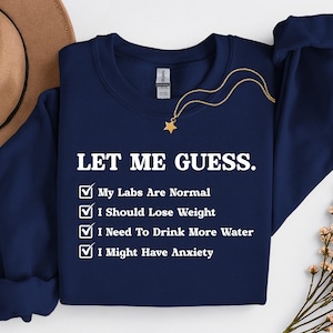 May include: Navy blue sweatshirt with the text "LET ME GUESS." and a list of relatable statements: "My Labs Are Normal," "I Should Lose Weight," "I Need To Drink More Water," and "I Might Have Anxiety." A gold necklace with a star pendant is visible.