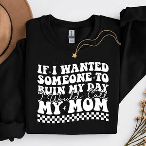 Może przedstawiać: Czarna bluza z okrągłym dekoltem z białym napisem "IF I WANTED SOMEONE TO RUIN MY DAY I Would Call MY MOM" w stylu retro. Widoczny jest również złoty naszyjnik z zawieszką w kształcie gwiazdy i brązowy kapelusz.