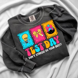 Pu&ograve; includere: Felpa grigio scuro con le parole "TEST DAY" in lettere colorate. La maglietta presenta tre grafiche quadrate con una lampadina, un fiocco e un becher scientifico. Il testo "DON'T STRESS! DO YOUR BEST!" &egrave; stampato anche sulla maglietta.