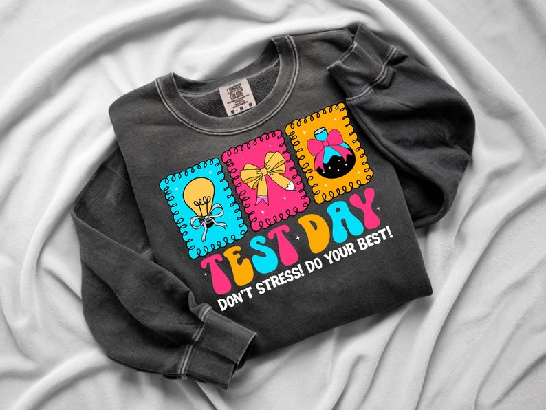 Pu&ograve; includere: Felpa grigio scuro con le parole "TEST DAY" in lettere colorate. La maglietta presenta tre grafiche quadrate con una lampadina, un fiocco e un becher scientifico. Il testo "DON'T STRESS! DO YOUR BEST!" &egrave; stampato anche sulla maglietta.