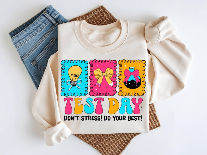 Pu&ograve; includere: Felpa color crema con grafica colorata che recita "TEST DAY" e "DON'T STRESS! DO YOUR BEST!". La grafica include una lampadina, un fiocco e un pallone. La felpa &egrave; abbinata a jeans blu.