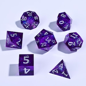 May include: A collection of seven polyhedral dice in various shapes, including a d20, d12, d100, d8, d6, d4, and a cube. The dice are a translucent purple with a galaxy-like interior and silver numbering. Ideal for tabletop gaming.