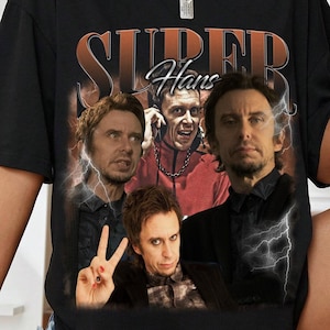 May include: Black t-shirt featuring a graphic design with the text "SUPER Hans" in orange and white. The design includes images of three men. The shirt is a casual, oversized style.