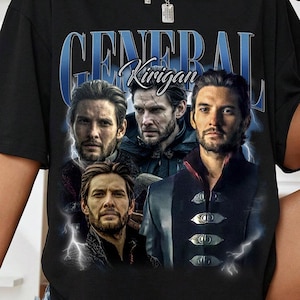 May include: Black t-shirt featuring a graphic of a man with the text "GENERAL Kirigan" in blue. The graphic includes multiple images of the man with lightning bolt accents. The shirt is a crew neck style.